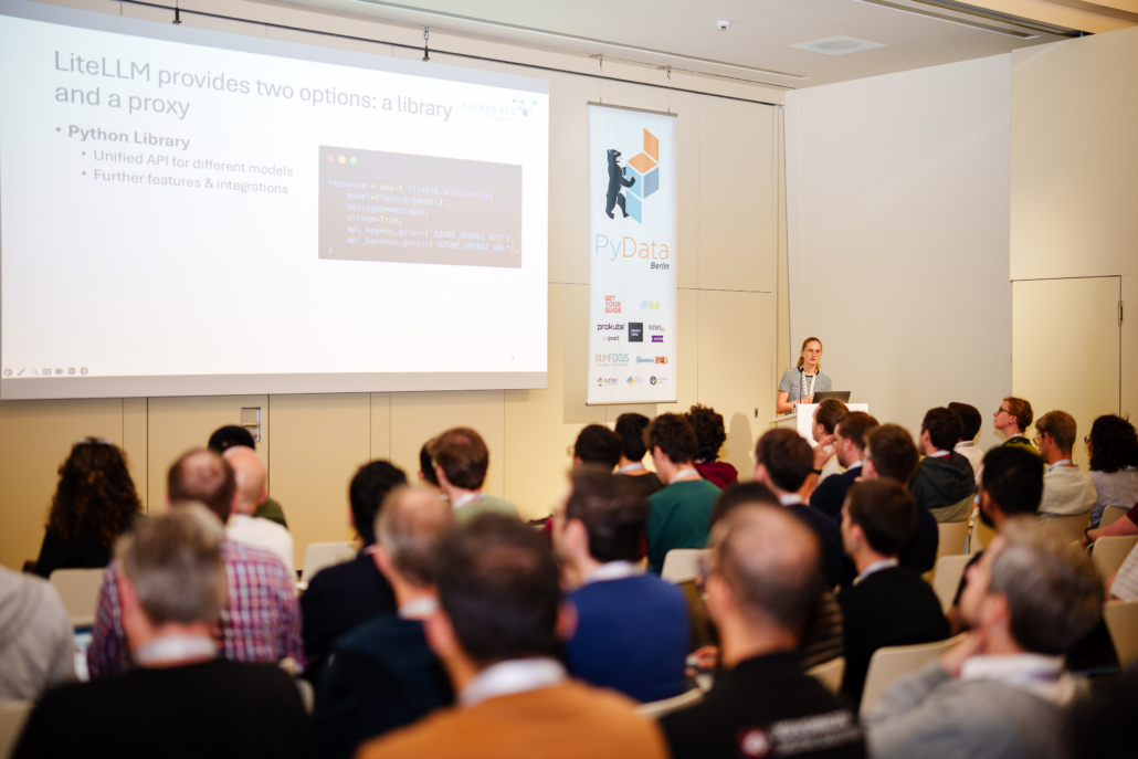 Eine Präsentation mit dem Titel 'LiteLLM provides two options: a library and a proxy' auf einer Leinwand. Alina Dallmann spricht vor einem Publikum, das aus vielen Personen besteht, die auf Stühlen sitzen. Auf dem Bildschirm sind Codebeispiele in einer Python-Bibliothek zu sehen.