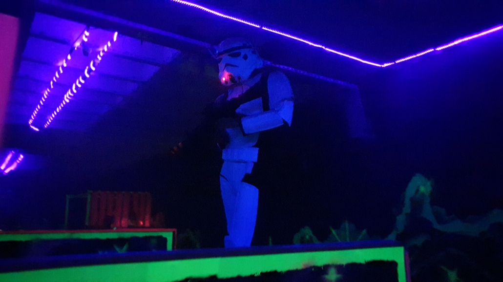 Figur in Stormtrooper-Kostüm mit Waffe in dunklem Raum mit violettem Neonlicht und grünen Leuchtflächen.