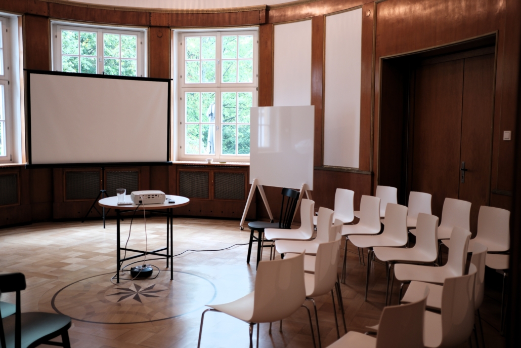 Leerer Seminarraum mit Stuhlreihen, rundem Tisch mit Projektor und Leinwand vor großen Fenstern.