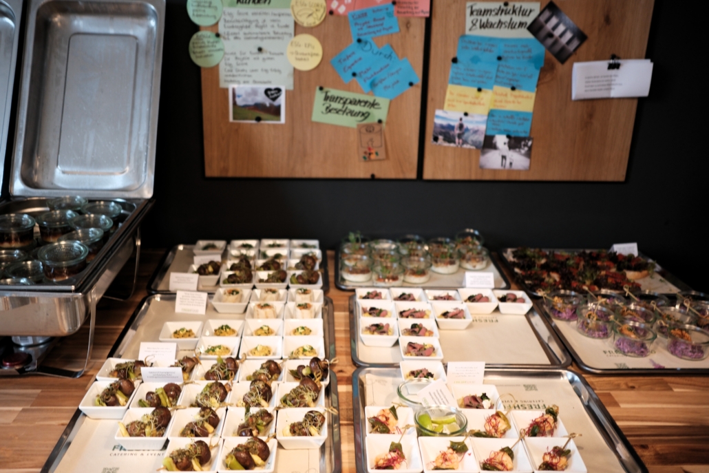 Buffet mit kleinen Portionen verschiedener Speisen in weißen Schalen und Gläsern auf Holzplatte, im Hintergrund Pinnwand mit Zetteln.