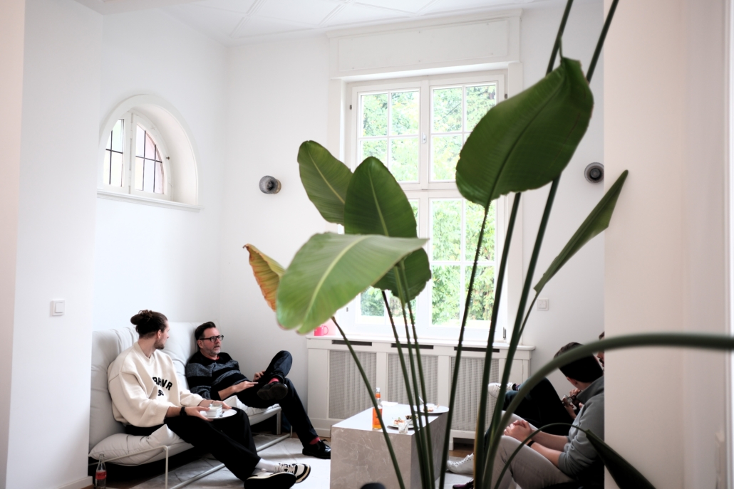 Mehrere Personen sitzen in einem hellen Raum mit großen Fenstern und weißen Wänden, im Vordergrund große grüne Blätter einer Pflanze.