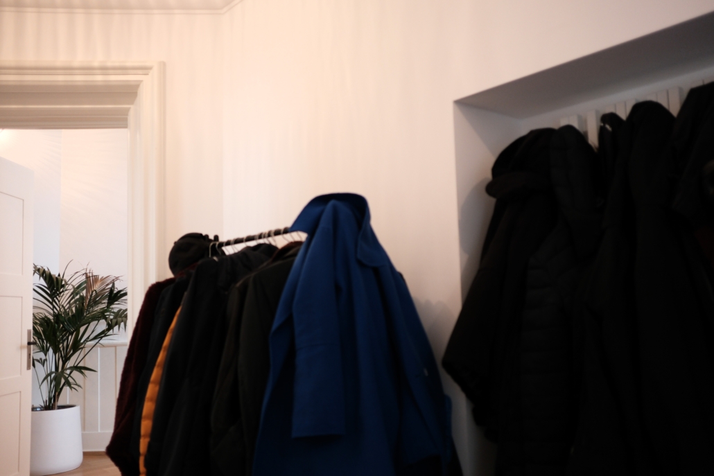 Mehrere Mäntel und Jacken hängen an einer Garderobe in einem Flur, links eine Pflanze in einem weißen Topf.