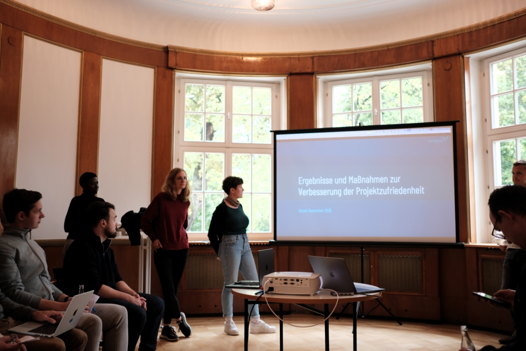 Mehrere Personen in einem Raum mit großen Fenstern, zwei stehen vor einer Leinwand mit der Projektion 'Ergebnisse und Maßnahmen zur Verbesserung der Projektzufriedenheit'.