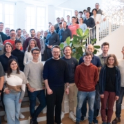 Gruppe von Personen steht auf und vor einer Treppe in einem hellen Raum mit großen Fenstern und einer runden Deckenleuchte.