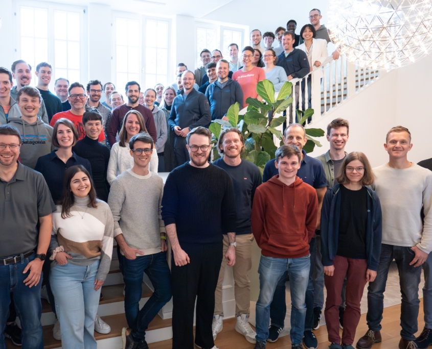 Gruppe von Personen steht auf und vor einer Treppe in einem hellen Raum mit großen Fenstern und einer runden Deckenleuchte.