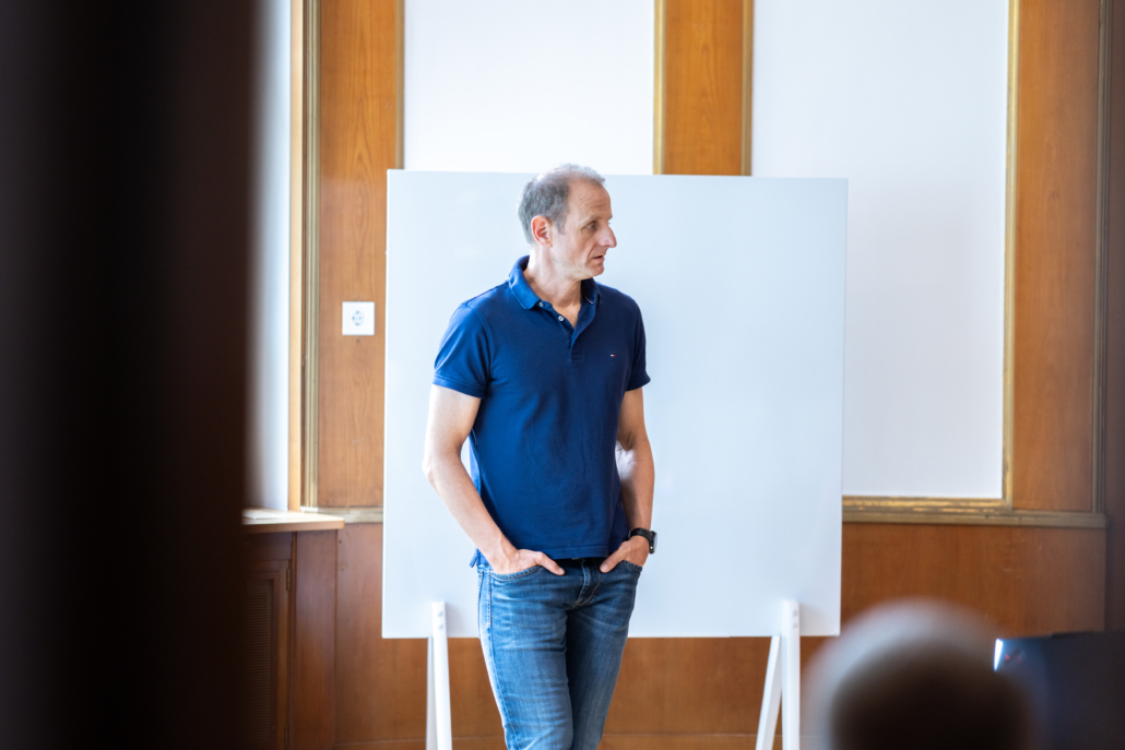 Mann in blauem Poloshirt und Jeans steht vor einem weißen Flipchart in einem Raum mit Holzvertäfelung.
