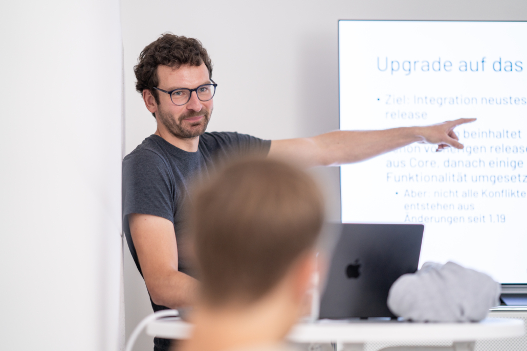 Person mit Brille und dunklem T-Shirt zeigt auf Präsentationsfolie mit Text 'Upgrade auf das ...'.