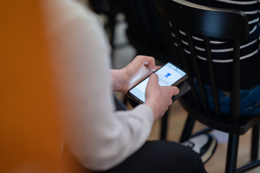 Person sitzt auf Stuhl und hält ein Smartphone in der Hand, trägt gestreiftes Oberteil und schwarze Hose.